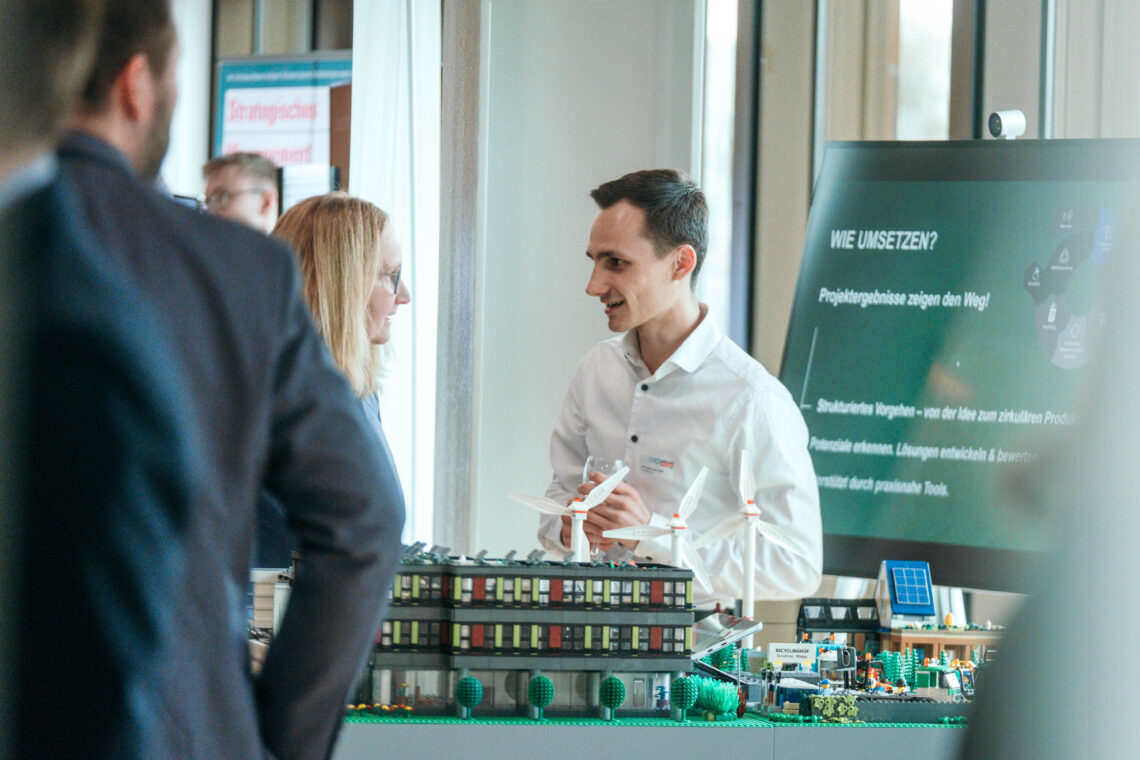 Personen im Gespräch vor dem ZirkuPro-Demonstrator, einem Modell aus Klemmbausteinen mit Gebäuden, Windrädern und Recyclingstation, das die Prinzipien der Kreislaufwirtschaft anschaulich visualisiert.